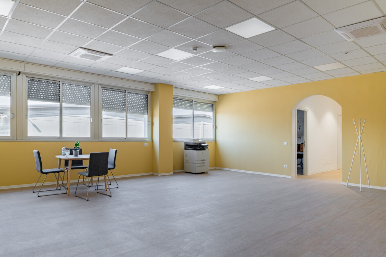 Ampia stanza d'ufficio con pareti gialle, grandi finestre, un tavolo con quattro sedie, una stampante e un appendiabiti.