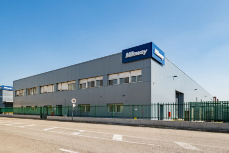 Un moderno edificio industriale con un'insegna Mileway, una facciata grigia, molteplici finestre e una recinzione verde sotto un cielo azzurro e limpido.