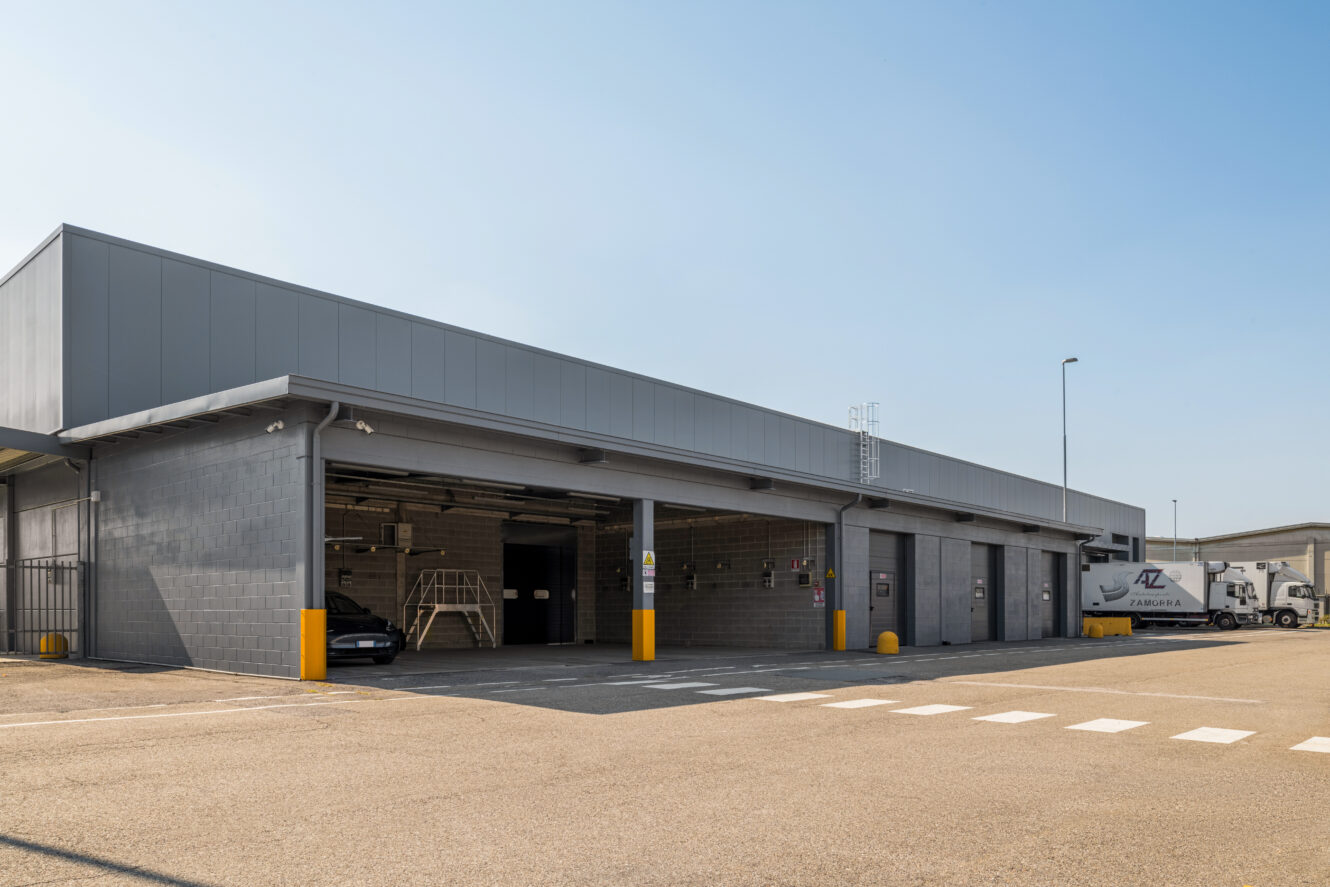 Un moderno edificio industriale con tre box aperti, un'auto nera all'interno e un camion di servizio parcheggiato nelle vicinanze in una giornata di sole.