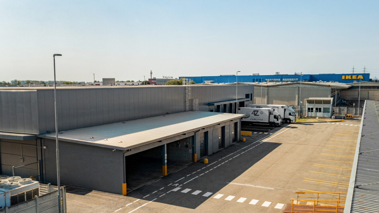 Un grande magazzino industriale con banchine di carico, un camion parcheggiato e un negozio IKEA sullo sfondo, sotto un cielo sereno.