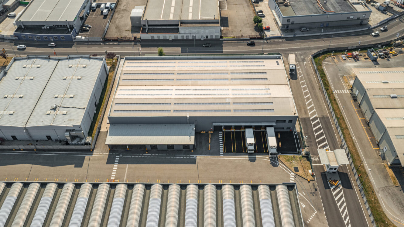 Vista aerea di un magazzino industriale con camion parcheggiati sulle banchine di carico, circondato da strade ed edifici adiacenti in un'area industriale.
