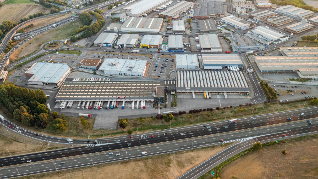 Veduta aerea di un complesso industriale con diversi magazzini, camion parcheggiati all'esterno e un'autostrada con veicoli in movimento in primo piano.