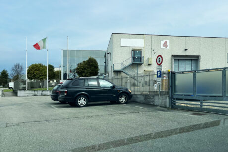 Un edificio industriale grigio con il numero 4, due auto parcheggiate, un cancello, cartelli di limite di velocità e di parcheggio e una bandiera italiana su un'asta.