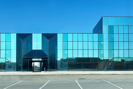 Un edificio moderno con finestre di vetro colorate di blu e un tetto piatto, visto dall'altra parte di un parcheggio vuoto sotto un cielo azzurro.
