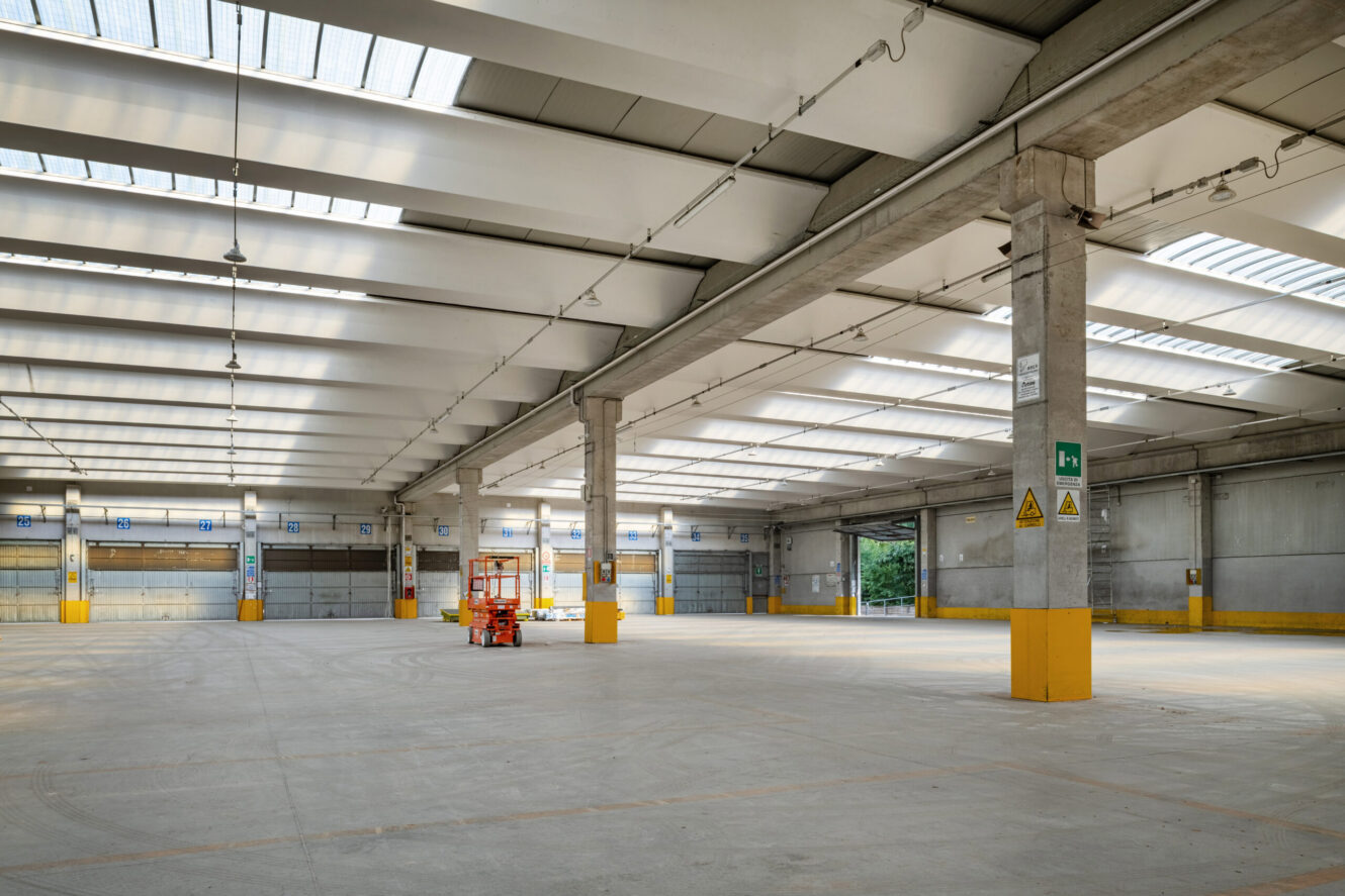 Interno di magazzino industriale spazioso e vuoto con soffitti alti, pavimenti in cemento, segnaletica di sicurezza gialla e un ascensore a forbice vicino al centro.