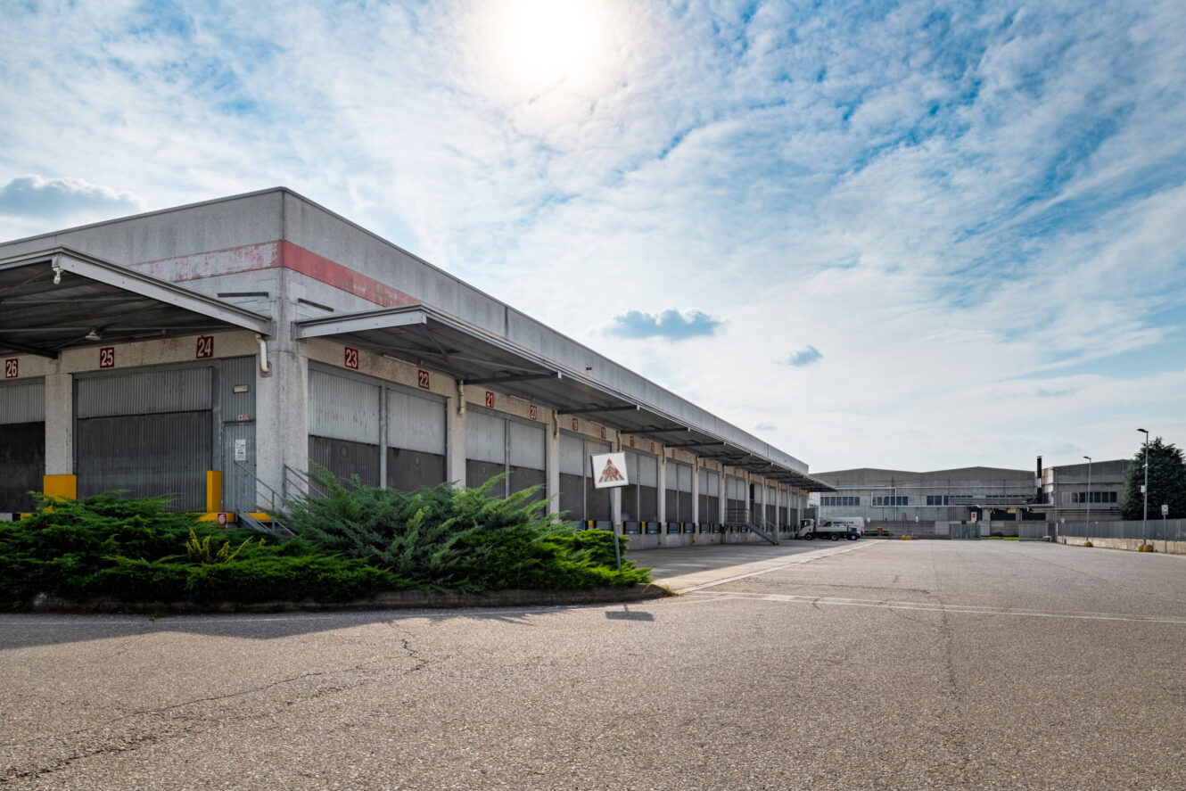 Grande edificio industriale vuoto con diverse banchine di carico e un lotto asfaltato sotto un cielo parzialmente nuvoloso.