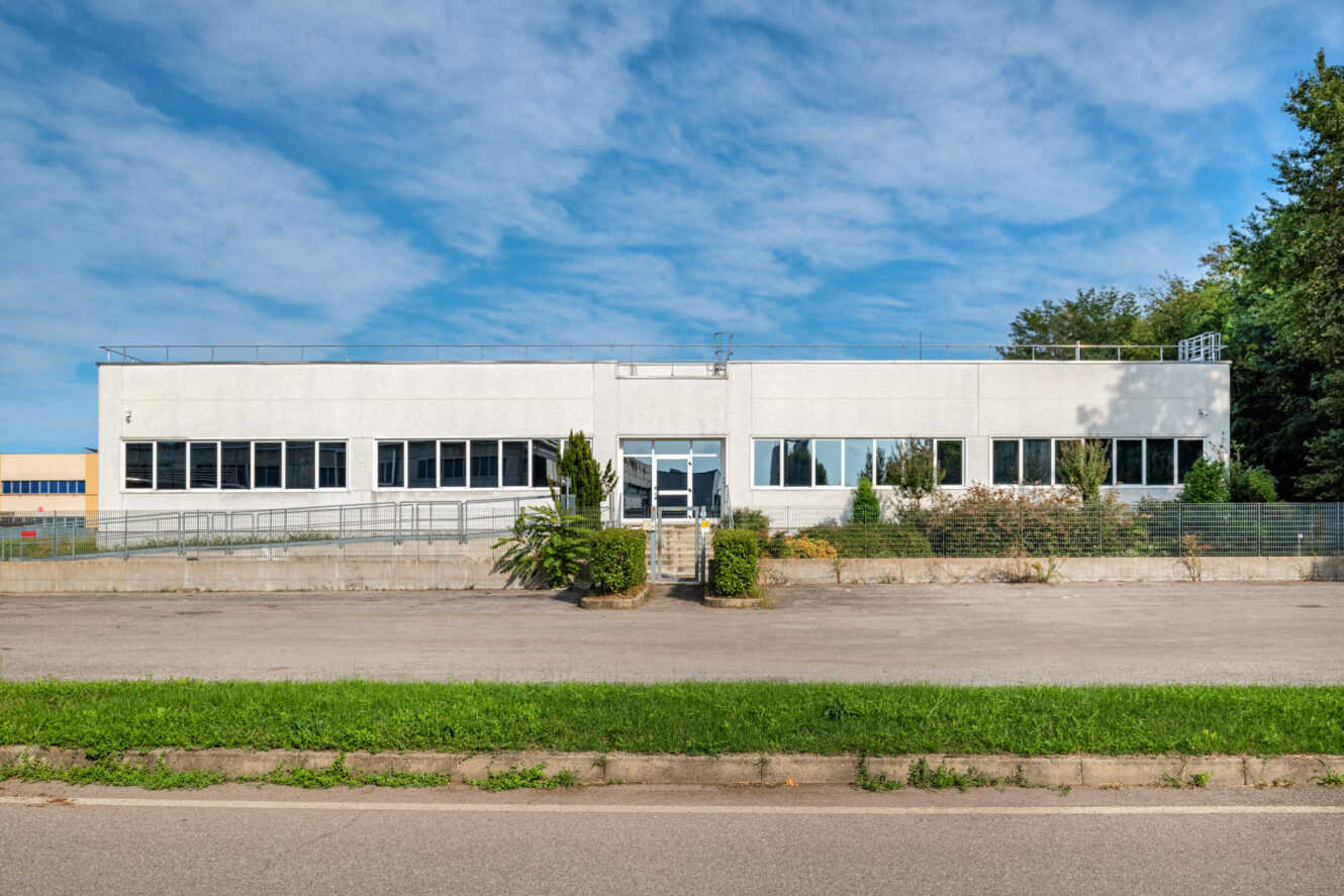 Un edificio commerciale o industriale rettangolare, bianco, a due piani, con grandi finestre, circondato dal verde, sotto un cielo azzurro.