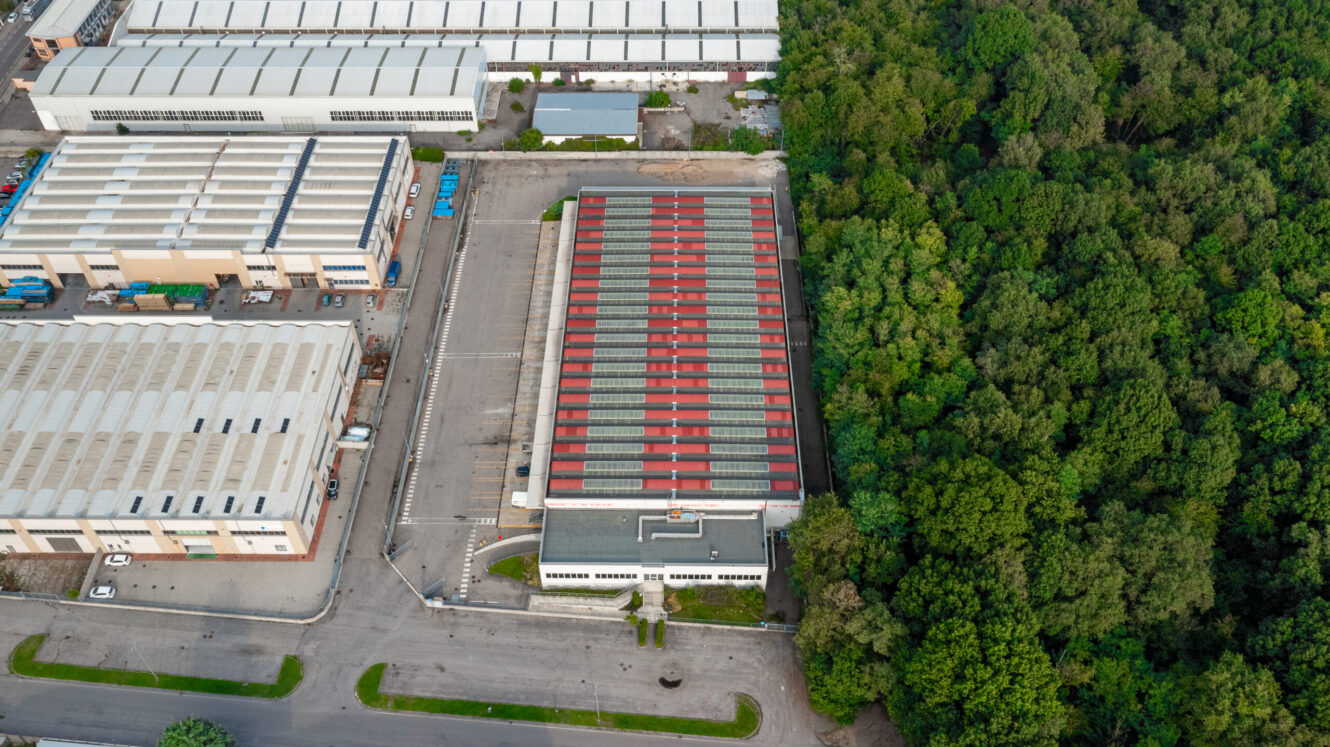 Vista aerea di un complesso di magazzini industriali vicino a una fitta area boschiva, con strade asfaltate e parcheggi visibili.