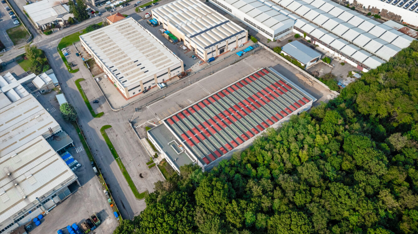 Veduta aerea di un'area industriale con più capannoni di grandi dimensioni, strade asfaltate, parcheggi e bosco verde adiacente.