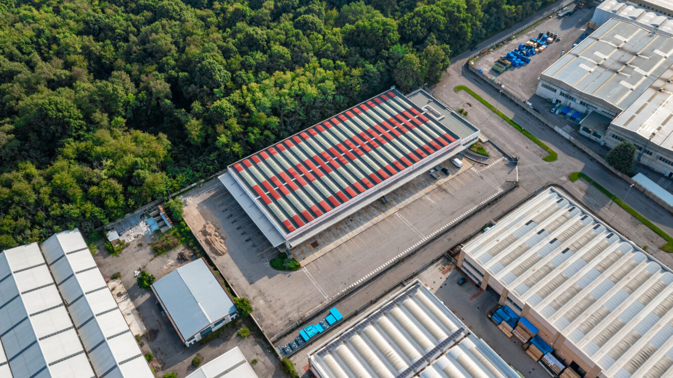 Veduta aerea di un grande magazzino industriale con tetto a strisce rosse, circondato da altri magazzini e da fitti alberi verdi.