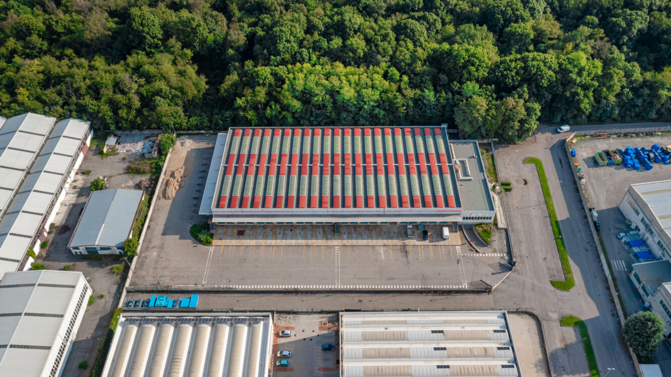 Veduta aerea di un grande magazzino con tetto a strisce rosse e grigie, circondato da strade, parcheggi, altri edifici industriali e fitti alberi verdi.