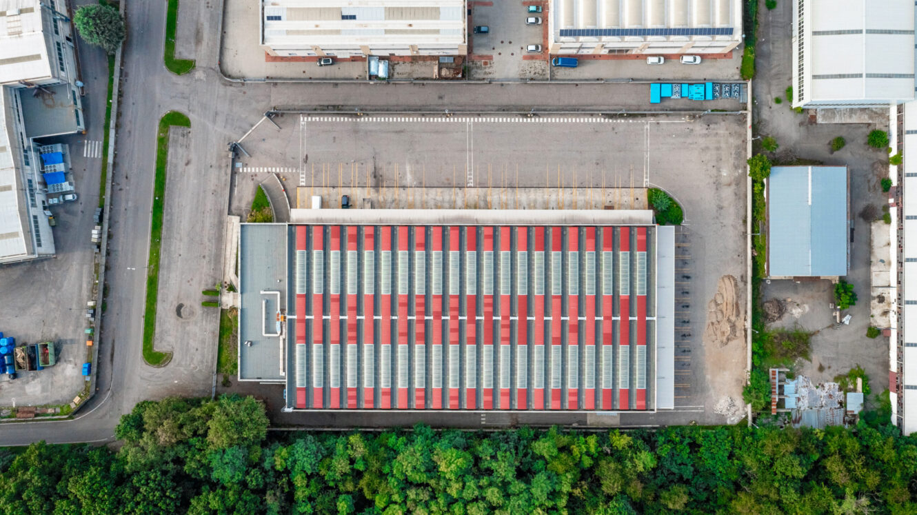 Veduta aerea di un grande magazzino industriale con tetto a strisce bianche e rosse, circondato da alberi, strade ed edifici adiacenti.