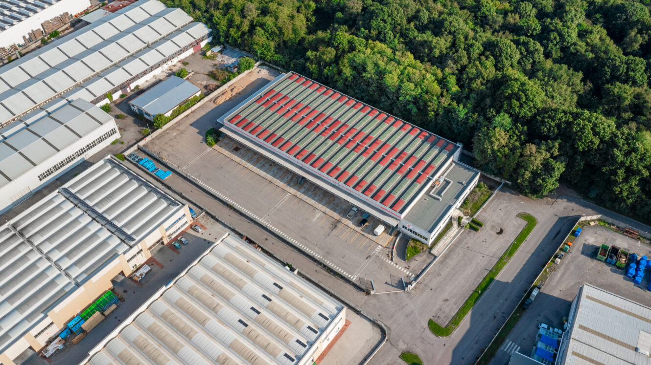 Veduta aerea di un grande capannone industriale con tetto rosso e bianco, circondato da aree di parcheggio, edifici adiacenti e un'area boschiva.