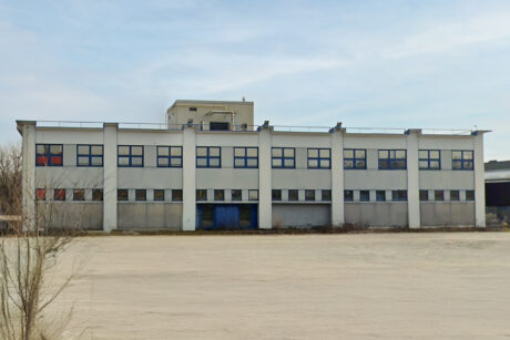 Un edificio industriale a due piani con molte finestre, un tetto piatto e una piccola struttura superiore, accanto a un lotto spoglio e vuoto sotto un cielo nuvoloso.