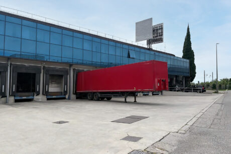 Un semirimorchio rosso è parcheggiato su una banchina di carico all'esterno di un moderno edificio di vetro in una giornata limpida.