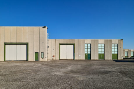 Grande magazzino industriale con pareti di cemento beige, porte multiple verdi e bianche e finestre alte, sullo sfondo di un cielo azzurro e di un terreno di ghiaia vuoto.
