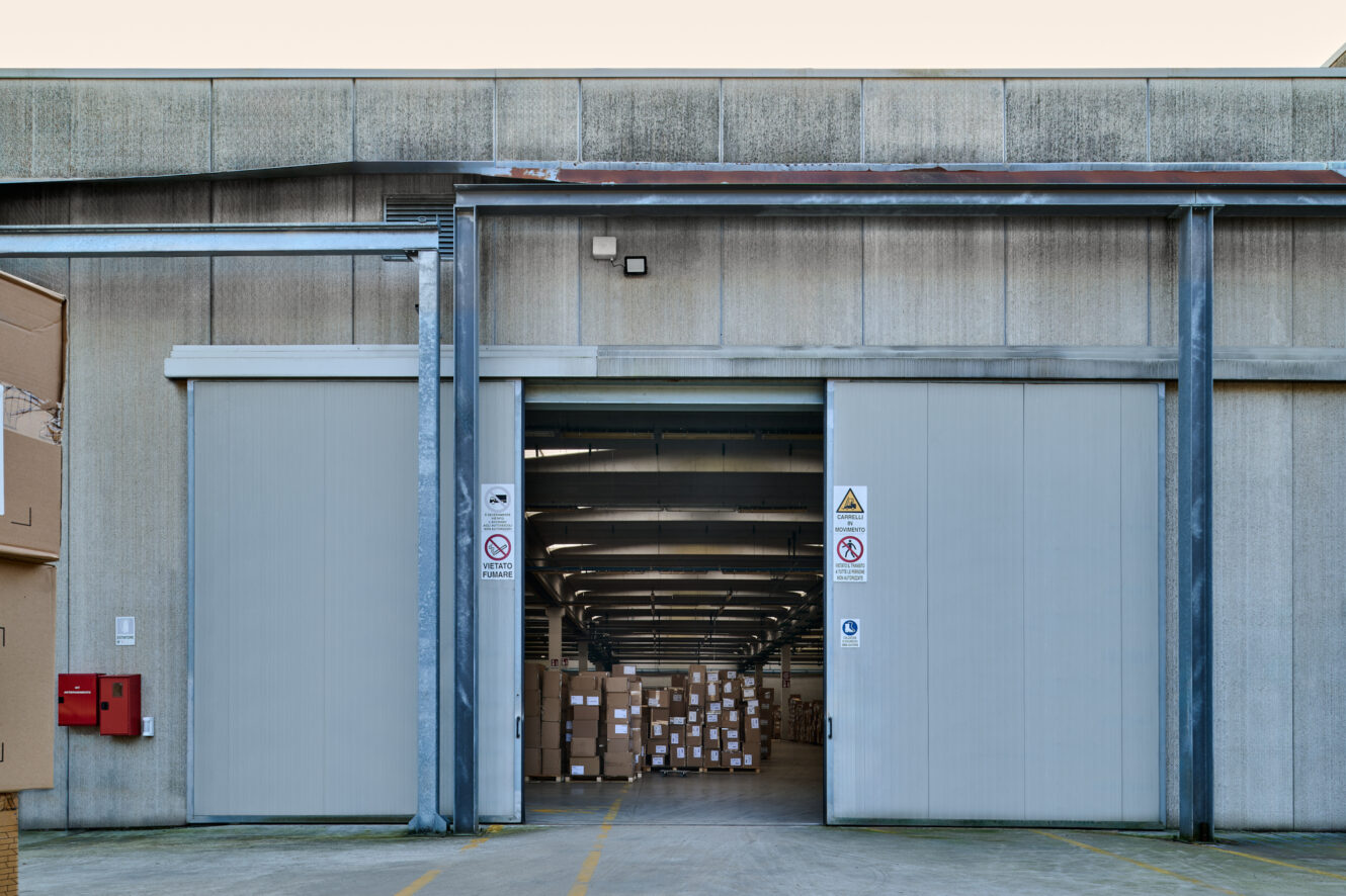 Magazzino con grandi porte scorrevoli aperte, che mostrano pile di scatole di cartone all'interno; cartelli di sicurezza e divieto di fumo affissi all'ingresso.