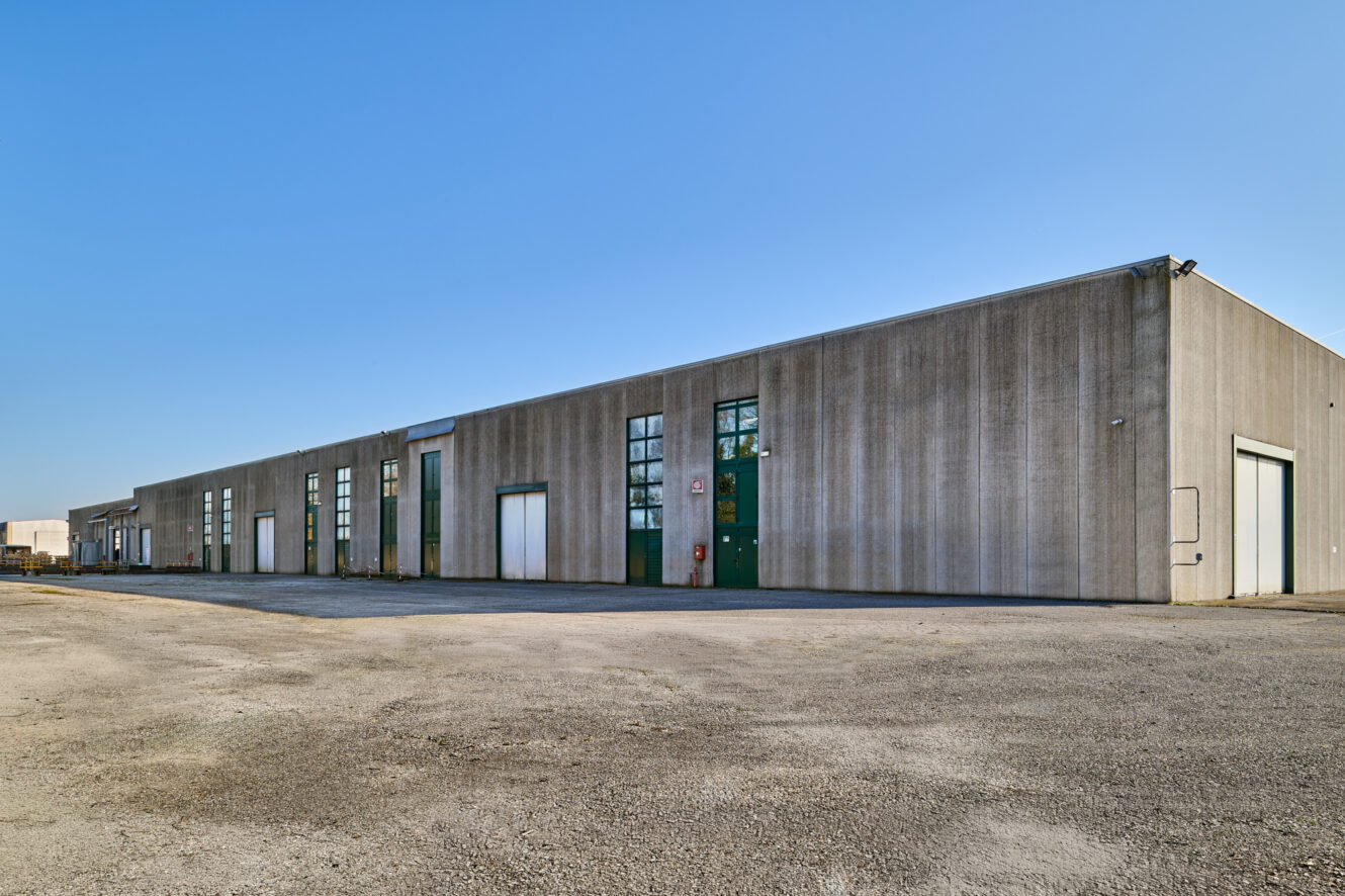 Un grande capannone industriale rettangolare con molteplici porte e finestre verdi, situato su un ampio terreno asfaltato e vuoto, sotto un cielo azzurro e limpido.
