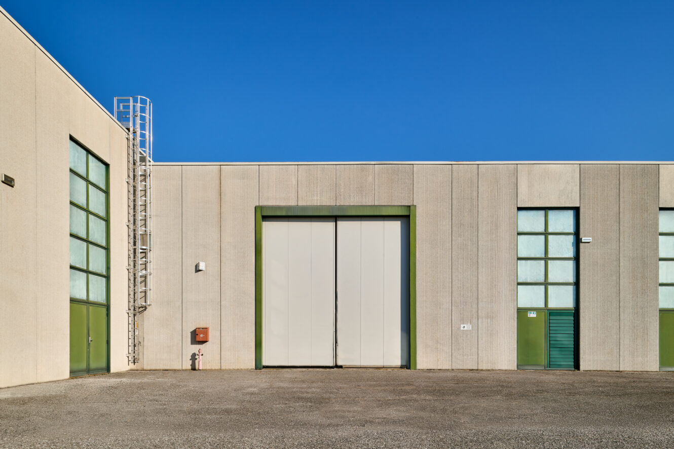 Esterno di un capannone industriale con un grande portone centrale, finestre e porte con cornice verde, una scala di metallo e un cielo azzurro.