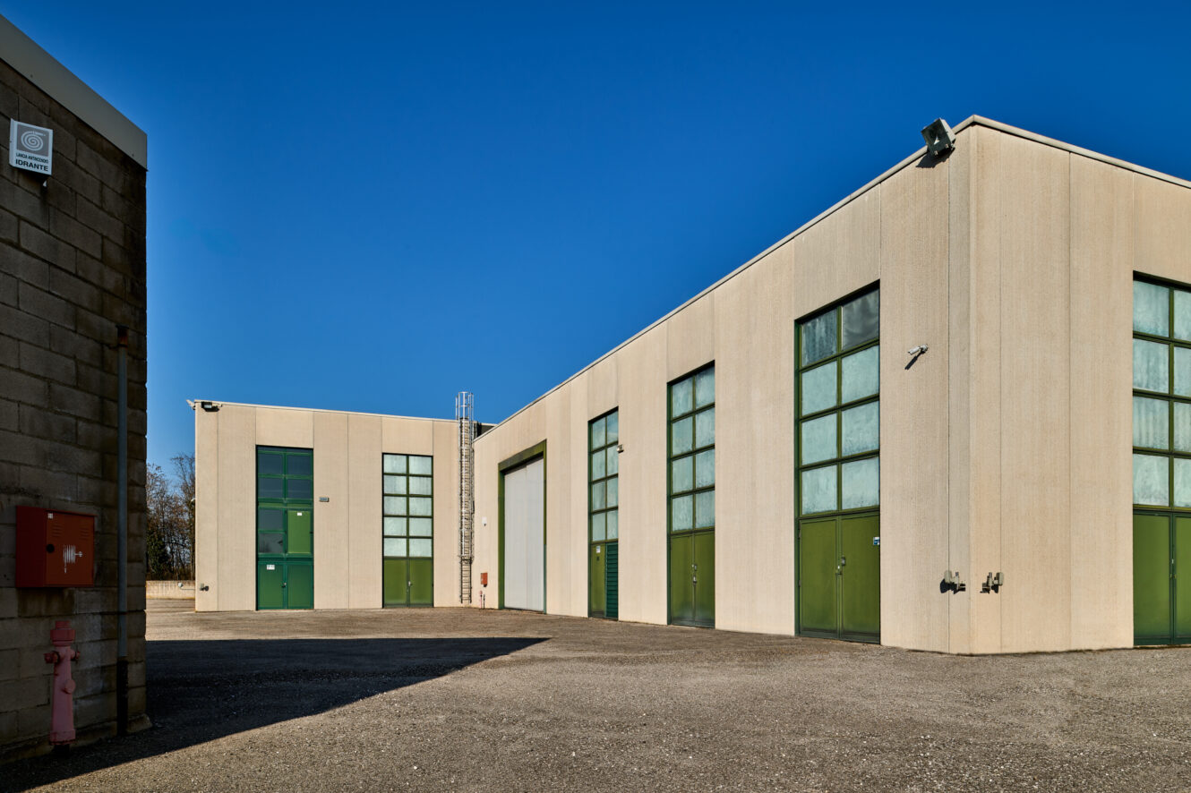 Due edifici industriali beige con grandi porte e finestre verdi sorgono su un terreno asfaltato sotto un cielo azzurro e limpido. In primo piano sono visibili un idrante rosso e una cassetta di allarme.