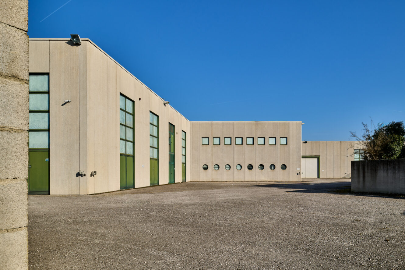 Un moderno edificio industriale con pareti beige, grandi porte verdi e finestre multiple, immerso in un cielo azzurro e con un terreno asfaltato vuoto davanti.