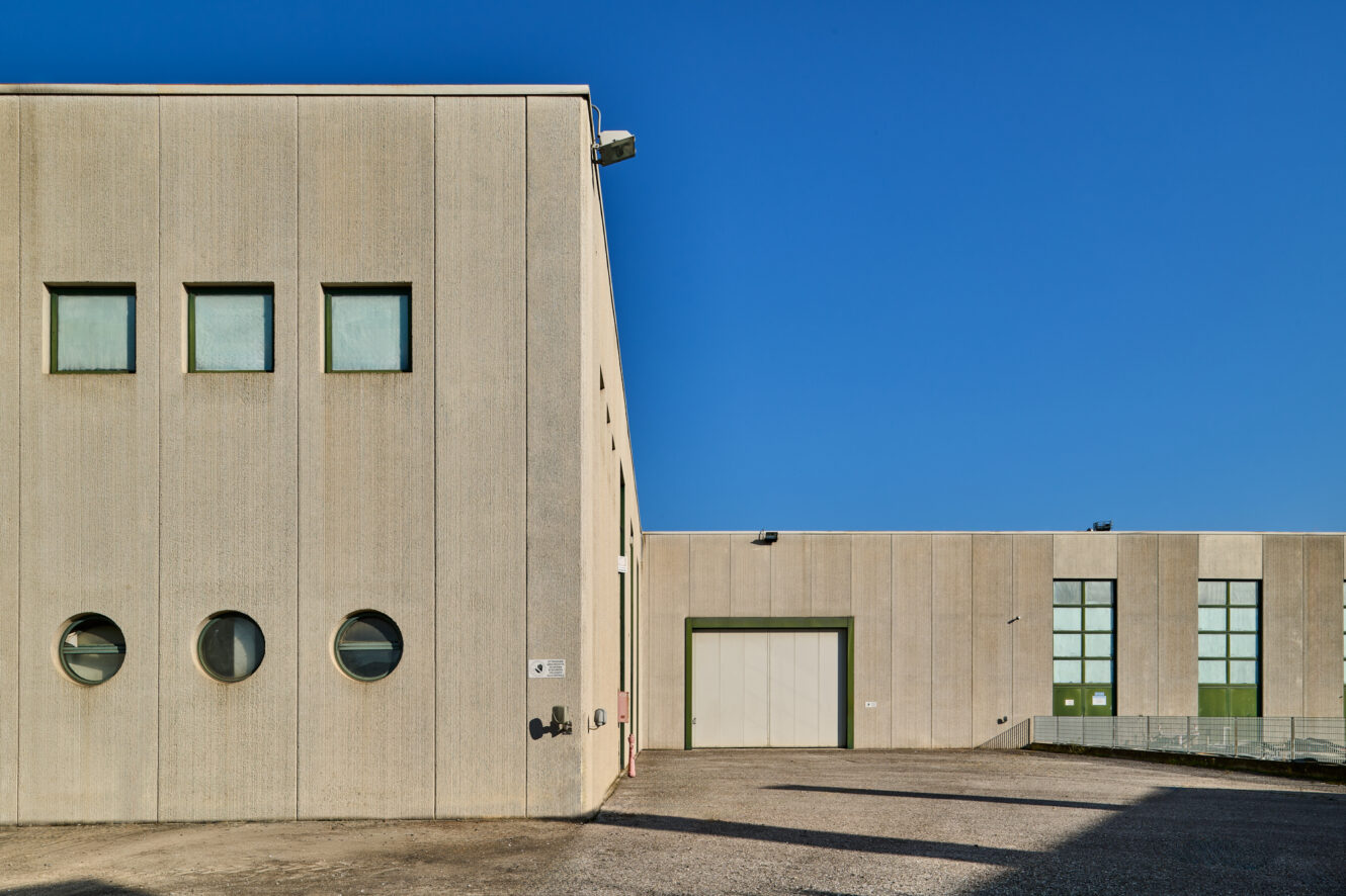 Edificio industriale con finestre rettangolari e circolari, pareti esterne beige e una grande porta di garage sotto un cielo azzurro.