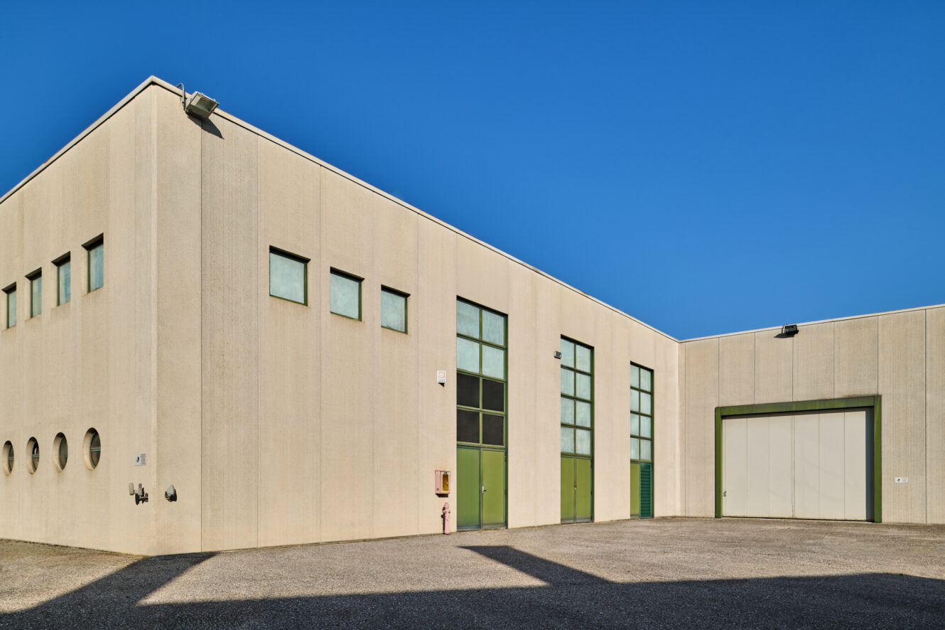 Un magazzino industriale beige con porte verdi e piccole finestre, fotografato sotto un cielo azzurro.