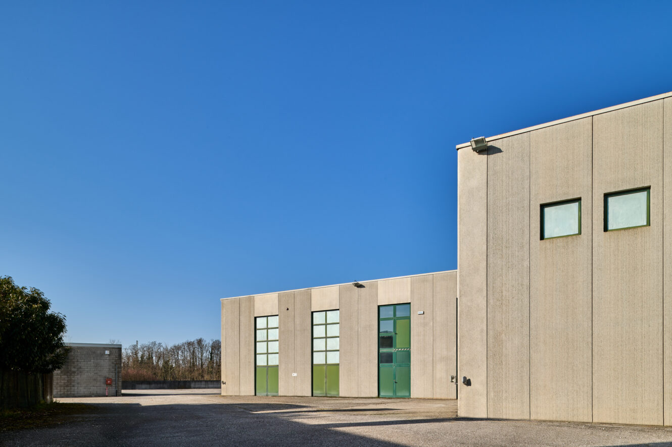 Moderni edifici industriali con pareti di cemento beige, grandi porte verdi e finestre rettangolari sotto un cielo azzurro.