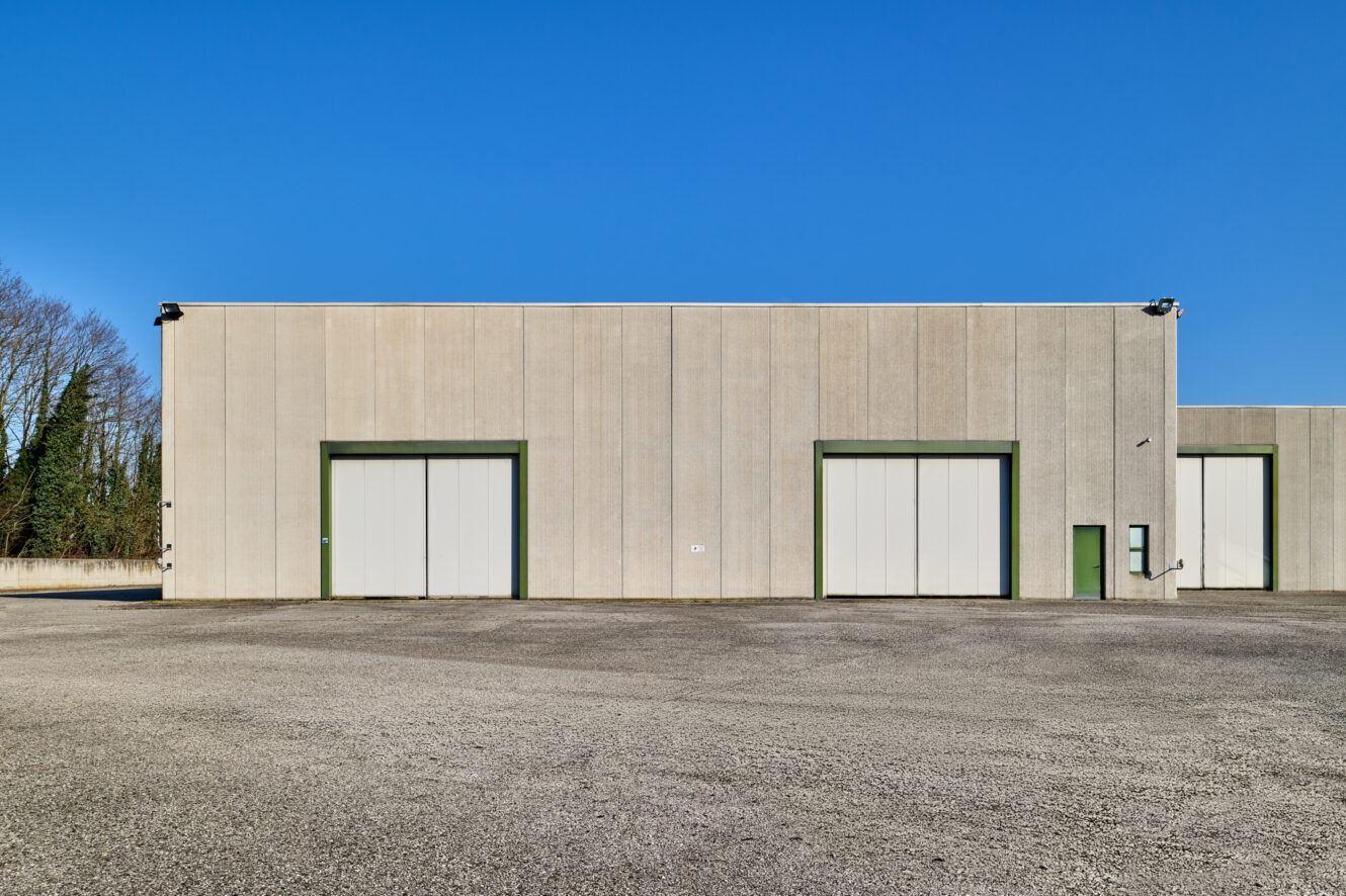 Un grande magazzino industriale grigio con due grandi porte chiuse e una porta verde più piccola, sullo sfondo di un cielo azzurro e di un terreno asfaltato vuoto.