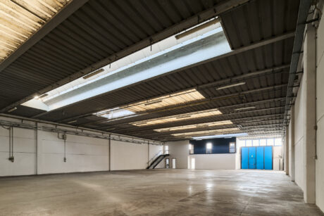 Interno di magazzino industriale vuoto con pavimento in cemento, soffitto alto, lucernari, pareti bianche, scale in metallo e grandi porte blu.