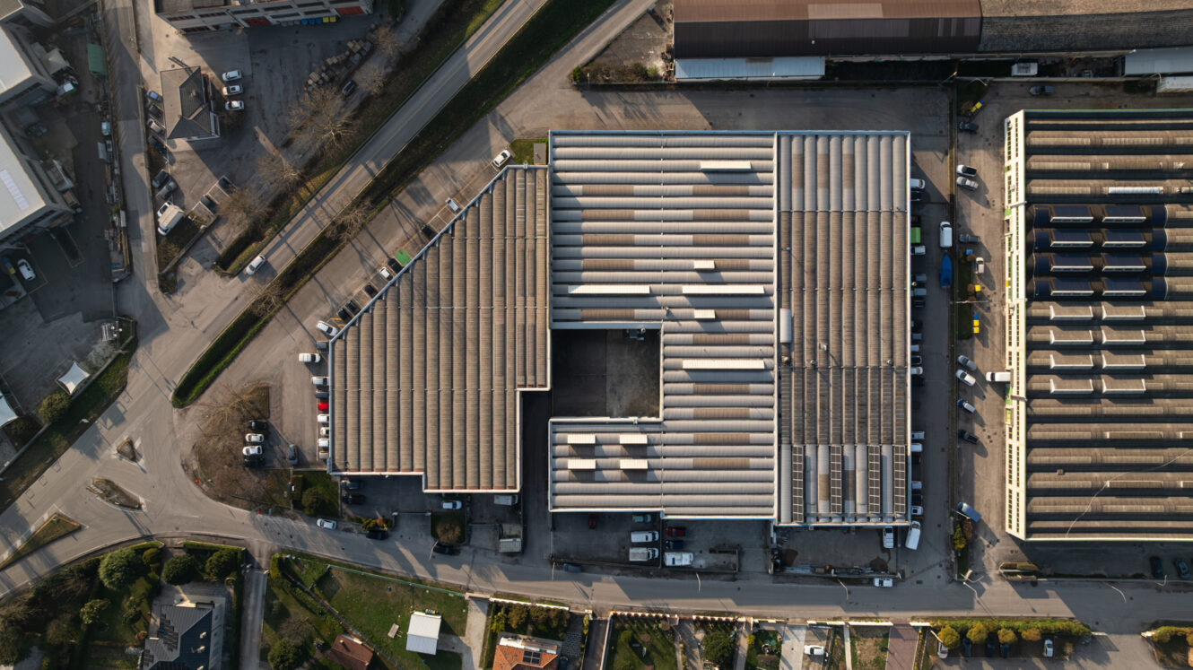Vista aerea di un grande edificio industriale a pianta rettangolare, circondato da strade, veicoli parcheggiati e strutture vicine.