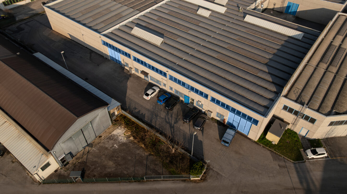 Vista aerea di un complesso di magazzini industriali con diverse auto e camion parcheggiati davanti a grandi porte blu.
