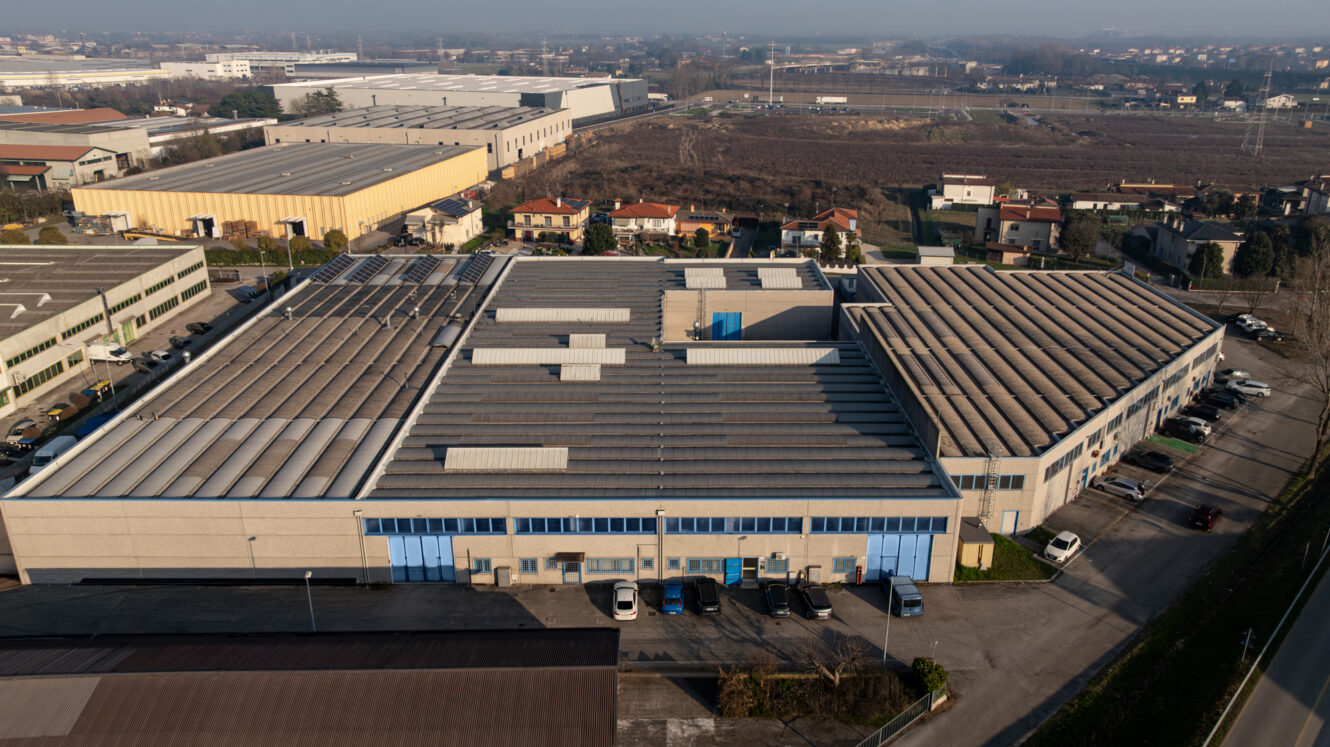Un grande complesso industriale con porte blu e lucernari sul tetto, circondato da edifici più piccoli, strade e terreni agricoli.