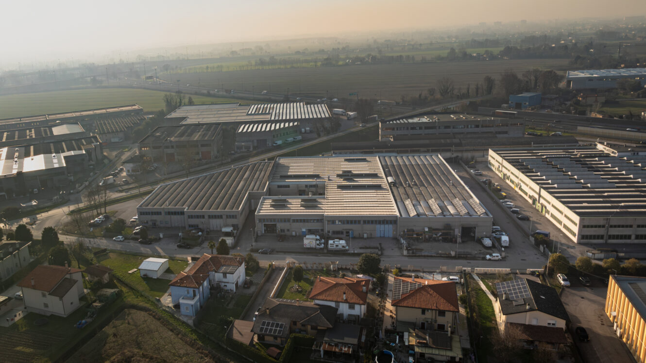 Veduta aerea di un complesso industriale con magazzini, camion e case residenziali delimitate da campi sotto un cielo nebuloso.