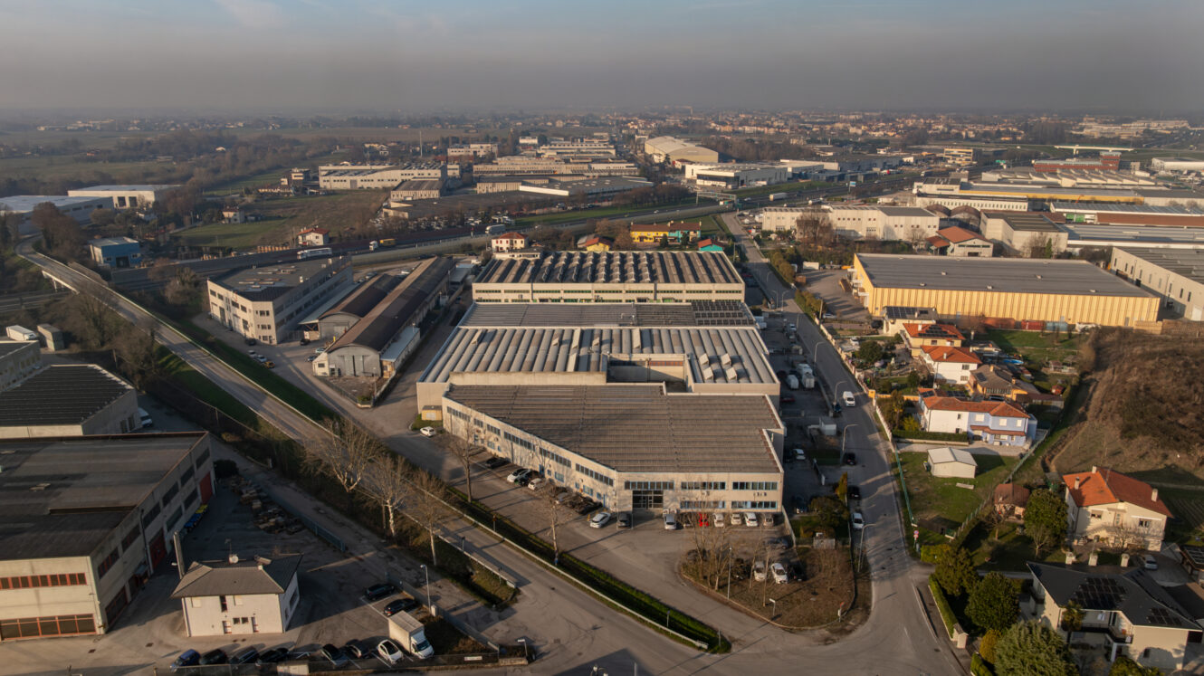Veduta aerea di un'area industriale con magazzini, fabbriche e case, circondata da strade e campi sotto un cielo nebuloso.