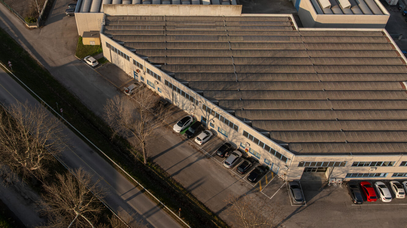 Vista aerea di un grande edificio industriale con auto parcheggiate all'esterno e alberi lungo la strada vicina.