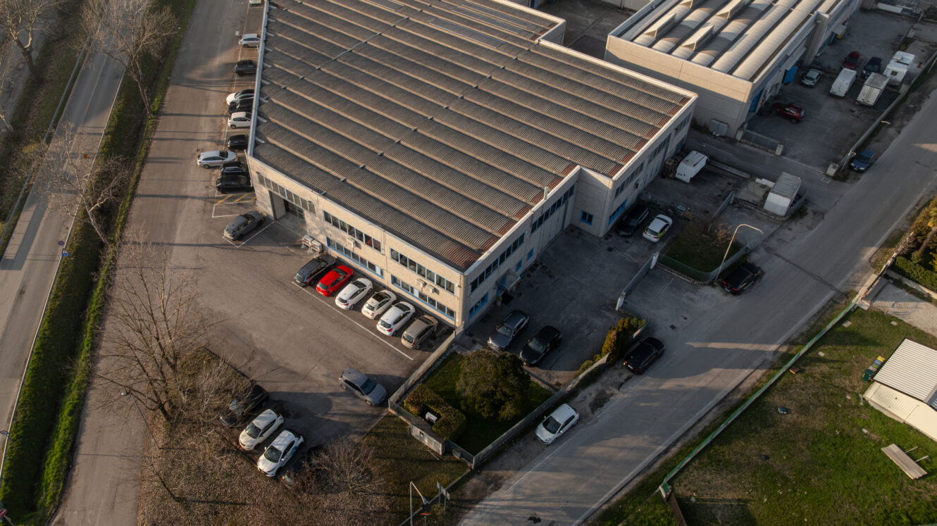 Vista aerea di un edificio industriale con un parcheggio; diverse auto sono parcheggiate intorno. Strade e verde circondano la proprietà.