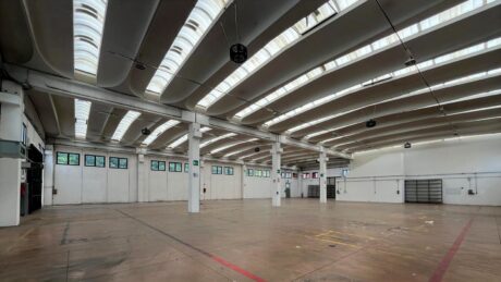 Grande magazzino vuoto con soffitti alti, travi a vista, lucernari, pavimento in cemento lucidato, piccole finestre e porte di garage.