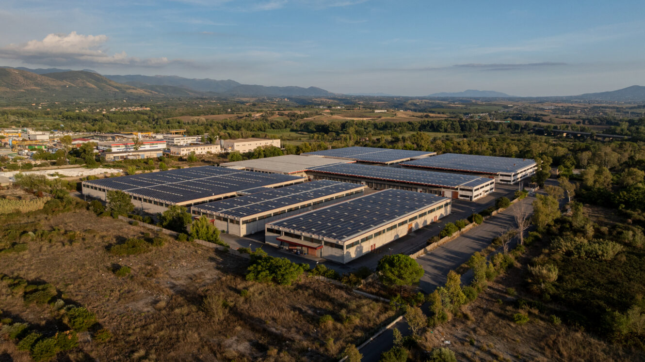 Vista aerea di un grande complesso industriale composto da diversi edifici, ciascuno con il tetto ricoperto di pannelli solari, circondato da campi e alberi sotto un cielo sereno.