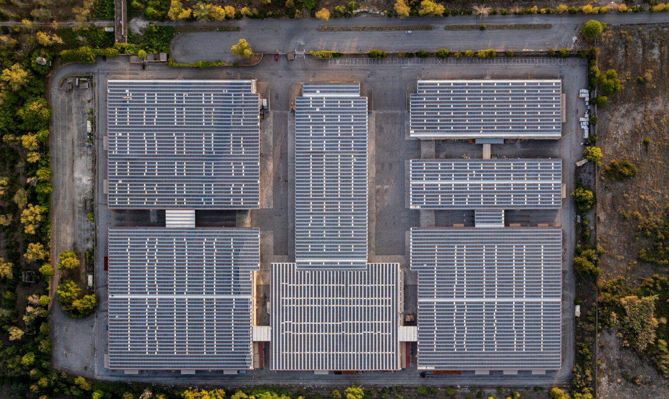 Vista aerea di un grande complesso industriale composto da diversi edifici con pannelli solari sui tetti, circondato da vegetazione e aree pavimentate.