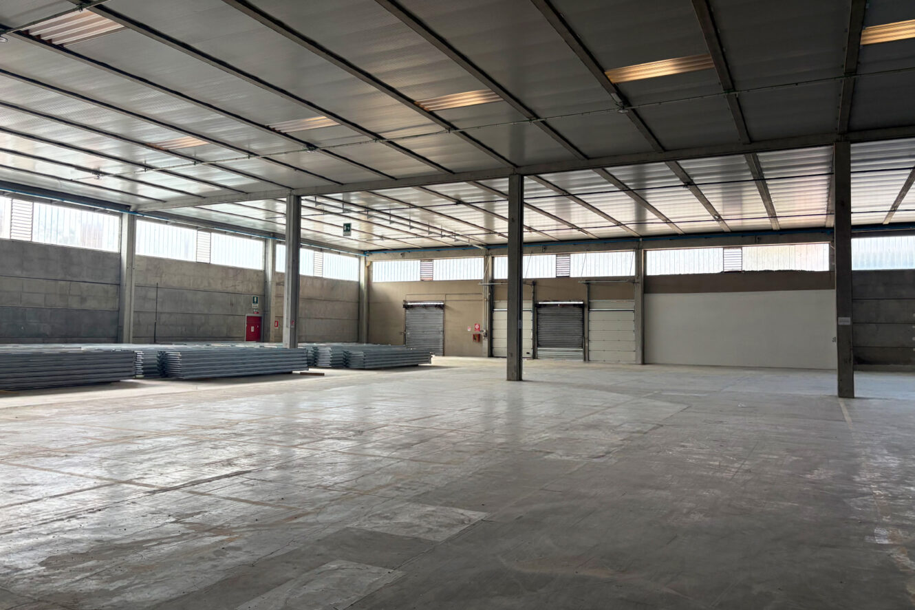 Interno di un magazzino vuoto con pavimenti in cemento, travi metalliche, materiali accatastati sulla sinistra e grandi porte industriali lungo la parete di fondo.