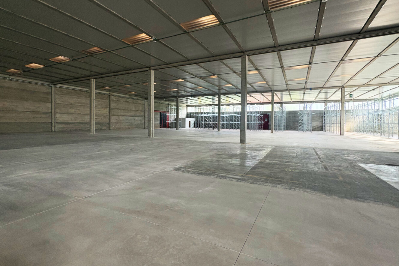 Interno di un capannone spazioso e vuoto, con pavimenti in cemento, soffitti alti, travi metalliche di sostegno e ampie finestre che lasciano entrare la luce naturale.