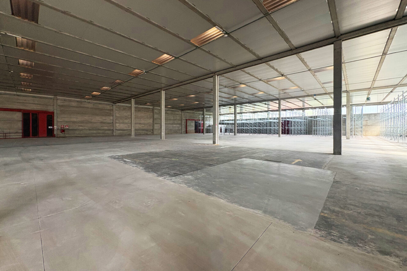 Interno di un capannone spazioso e vuoto, con pavimenti in cemento, soffitto a vista, colonne portanti e scaffalature lungo un lato. La luce naturale entra attraverso ampie finestre.