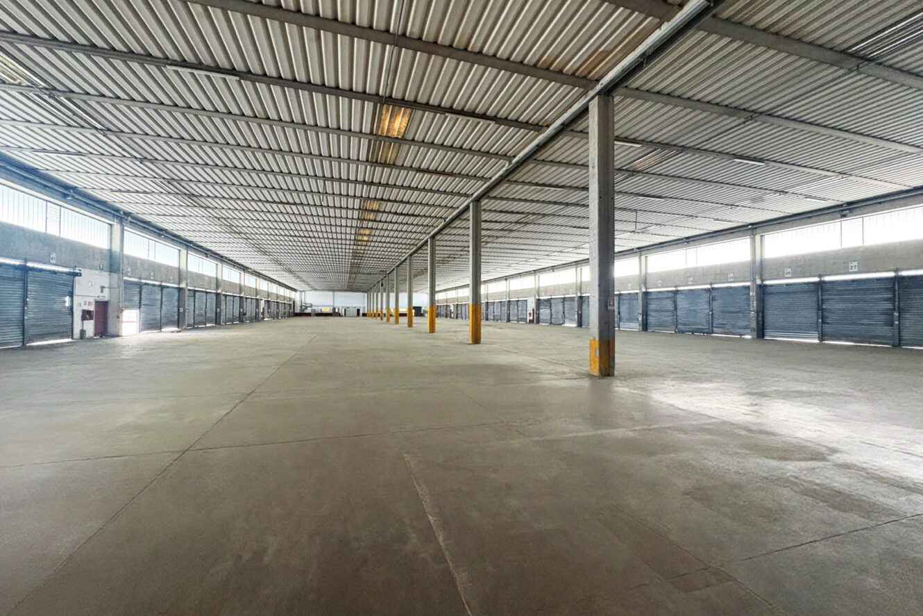 Ampio capannone industriale vuoto con pavimenti in cemento, pilastri metallici e file di porte di carico chiuse su entrambi i lati, sotto un tetto in lamiera ondulata.