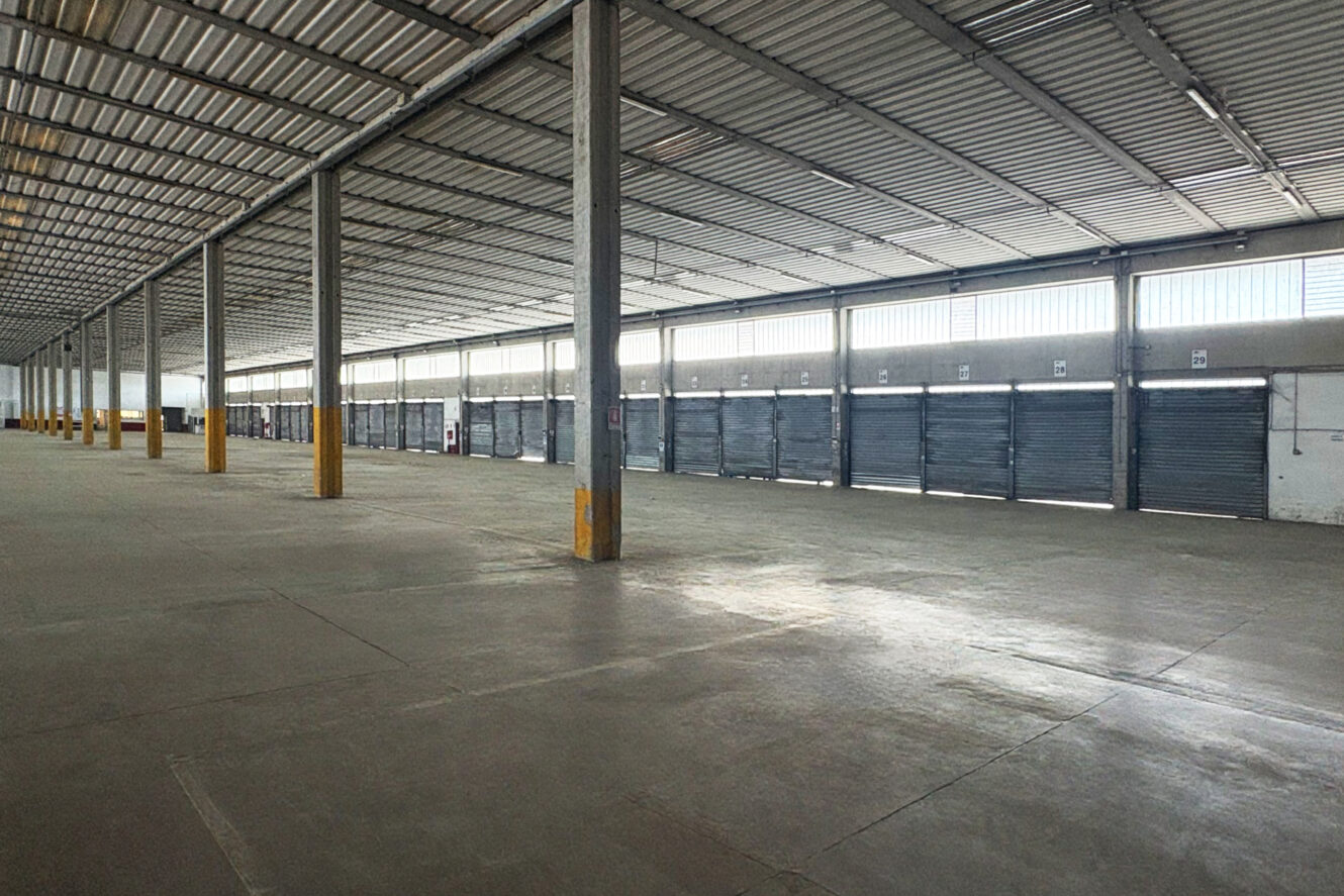 Interno di un grande magazzino vuoto, con pavimenti in cemento, pilastri metallici e diverse porte avvolgibili chiuse lungo un lato.