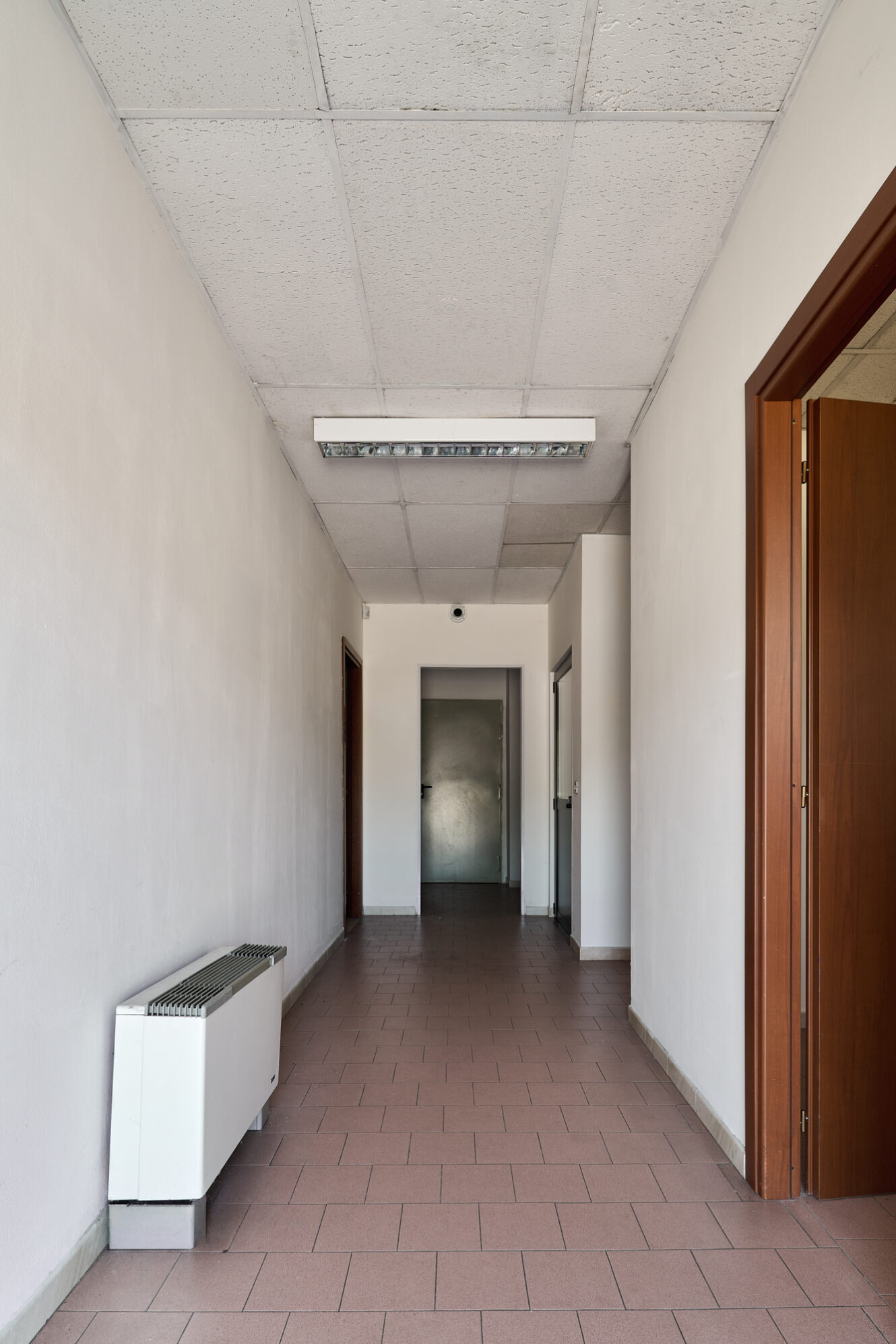 Un semplice corridoio con pareti bianche, pavimento piastrellato, un termosifone sulla sinistra e porte su entrambi i lati che conducono a una porta metallica chiusa in fondo.