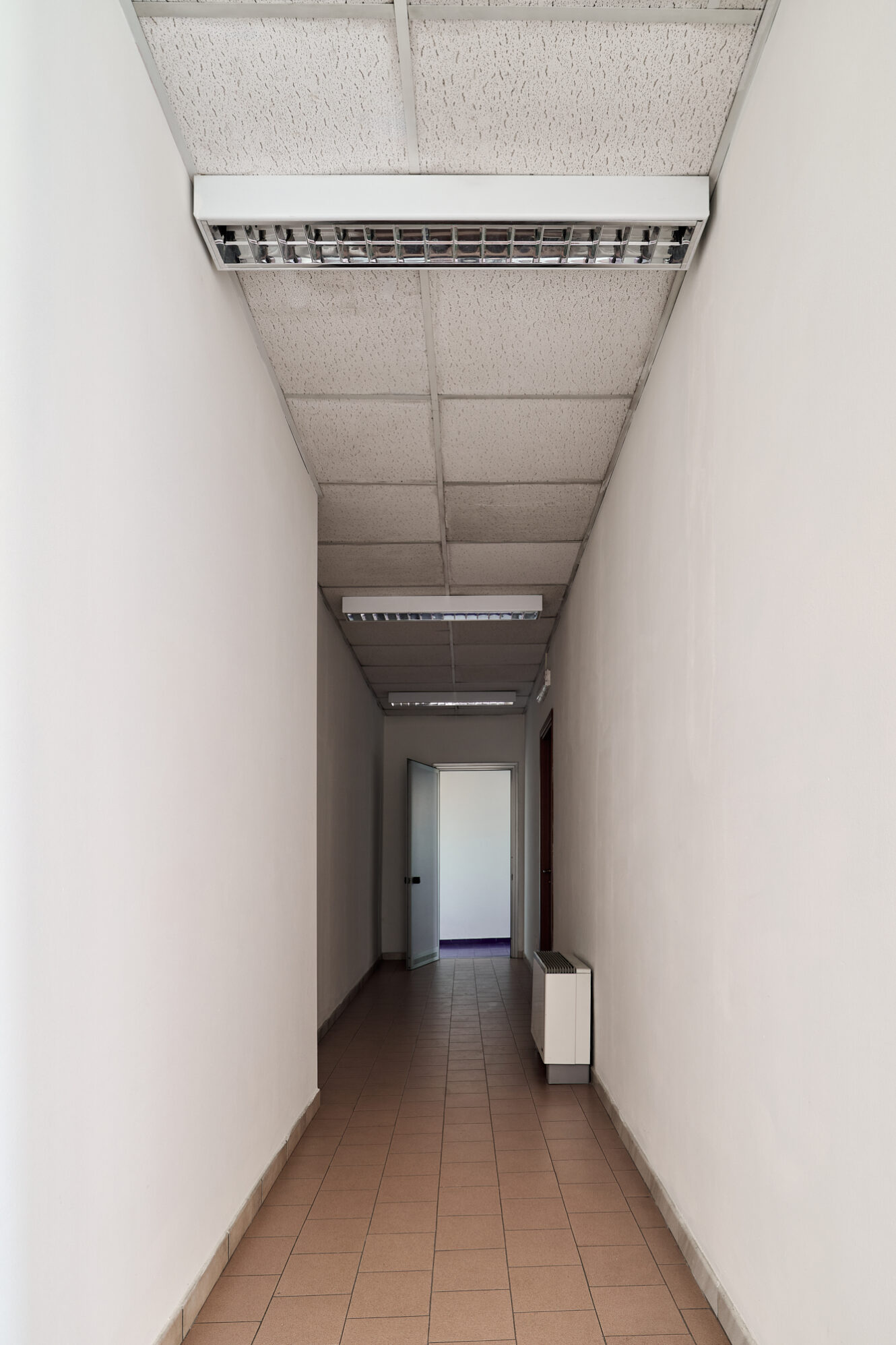 Un corridoio stretto con pareti bianche, pavimento piastrellato, luci a soffitto e, in fondo, una porta aperta che conduce a un’altra stanza.