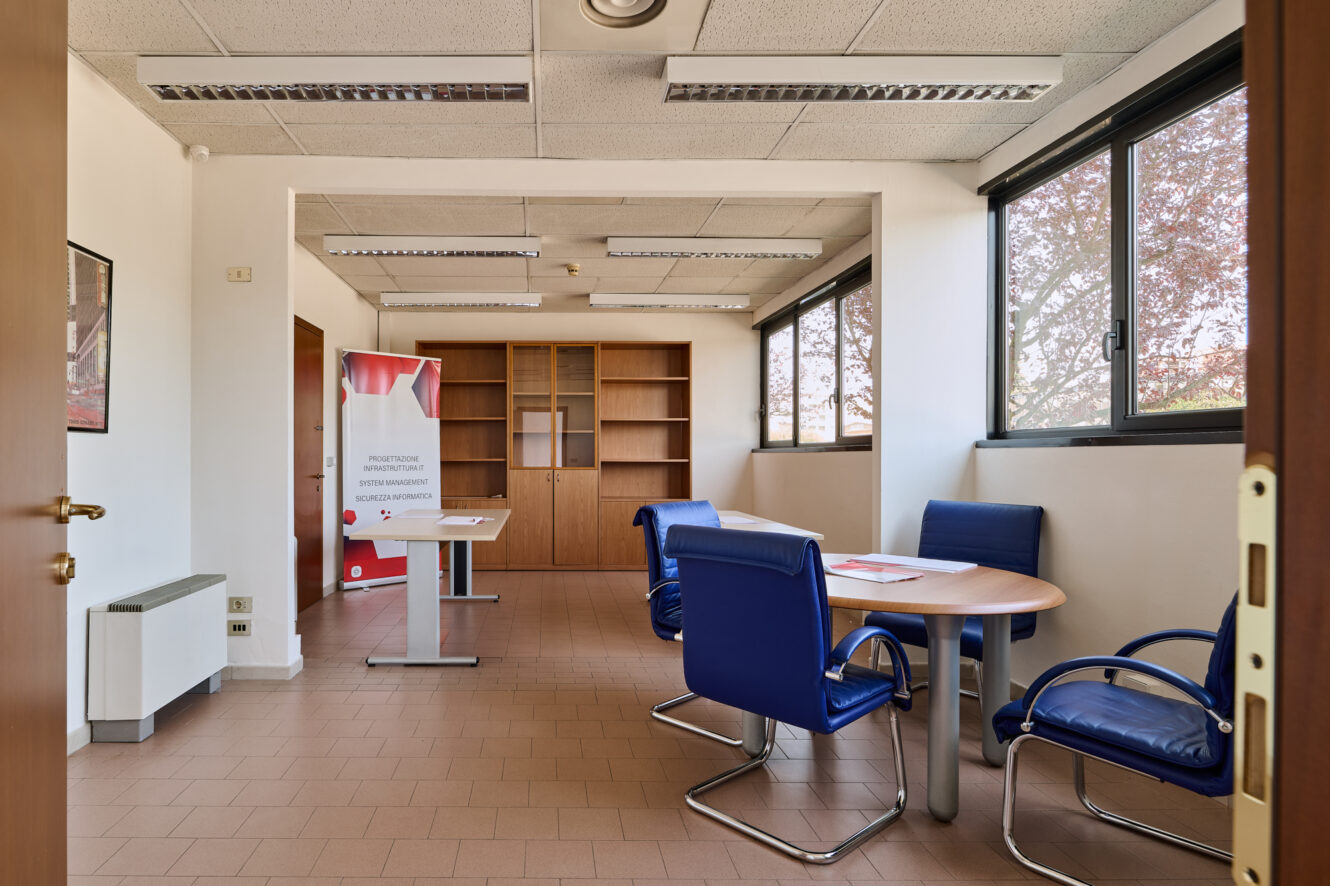 Stanza adibita a ufficio con sedie blu disposte attorno a un tavolo rotondo, una libreria in legno, una scrivania con uno striscione e ampie finestre che lasciano entrare la luce naturale.