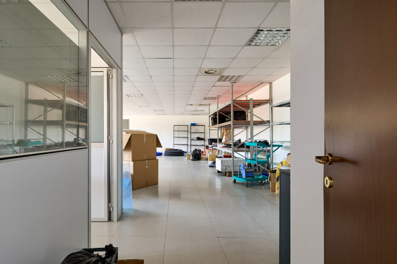 Vista di un magazzino con scaffali, scatole e materiale vario, ripresa attraverso una porta aperta, con pavimento piastrellato e luci fluorescenti a soffitto.