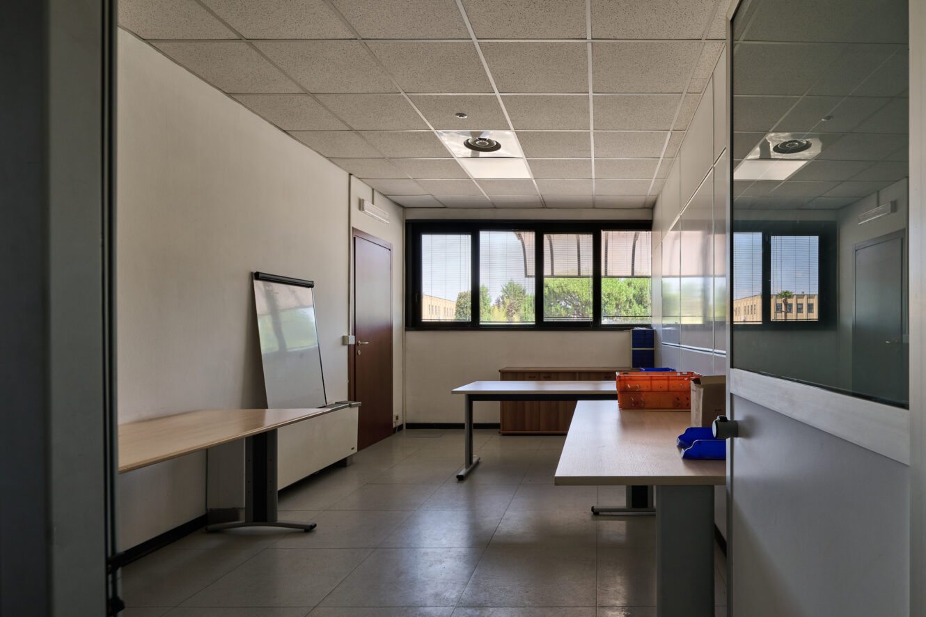 Un ufficio vuoto con due scrivanie, una lavagna bianca, ampie finestre e una porta. Su una delle scrivanie ci sono alcune scatole e materiale da ufficio. La luce naturale entra a fiotti dalle finestre.